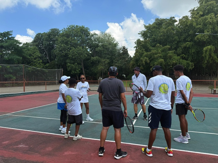Grupo de jugadores escucha indicaciones en una cancha de tenis.